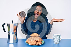 Beautiful african woman eating breakfast holding cholate donut celebrating achievement with happy smile and winner expression with