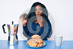 Beautiful african woman eating breakfast holding cholate donut covering mouth with hand, shocked and afraid for mistake
