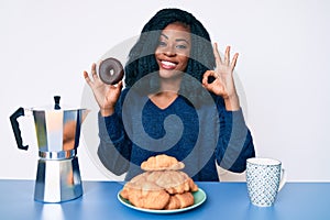 Beautiful african woman eating breakfast holding cholate donut doing ok sign with fingers, smiling friendly gesturing excellent