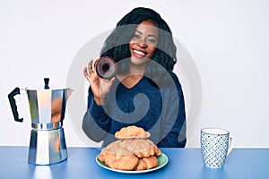 Beautiful african woman eating breakfast holding cholate donut looking positive and happy standing and smiling with a confident