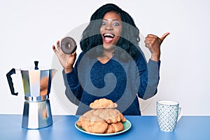 Beautiful african woman eating breakfast holding cholate donut pointing thumb up to the side smiling happy with open mouth