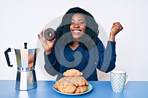 Beautiful african woman eating breakfast holding cholate donut screaming proud, celebrating victory and success very excited with