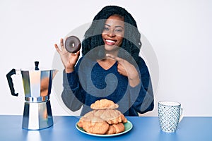 Beautiful african woman eating breakfast holding cholate donut smiling happy pointing with hand and finger