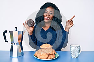 Beautiful african woman eating breakfast holding cholate donut smiling happy pointing with hand and finger to the side