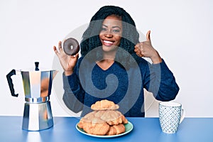 Beautiful african woman eating breakfast holding cholate donut smiling happy and positive, thumb up doing excellent and approval