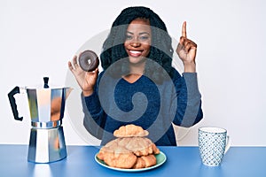 Beautiful african woman eating breakfast holding cholate donut smiling with an idea or question pointing finger with happy face,