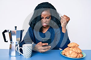 Beautiful african woman eating breakfast using smartphone annoyed and frustrated shouting with anger, yelling crazy with anger and