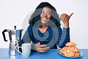 Beautiful african woman eating breakfast using smartphone pointing thumb up to the side smiling happy with open mouth