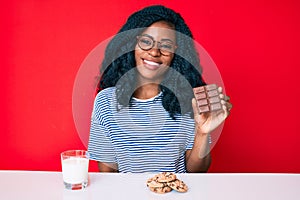 Beautiful african woman eating a chocolate bar and drinking glass of milk looking positive and happy standing and smiling with a