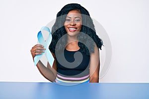 Beautiful african woman holding blue ribbon looking positive and happy standing and smiling with a confident smile showing teeth