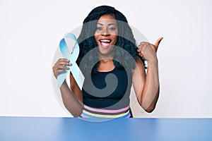 Beautiful african woman holding blue ribbon pointing thumb up to the side smiling happy with open mouth