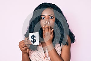 Beautiful african woman holding dollar symbol paper covering mouth with hand, shocked and afraid for mistake