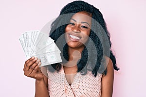 Beautiful african woman holding dollars looking positive and happy standing and smiling with a confident smile showing teeth