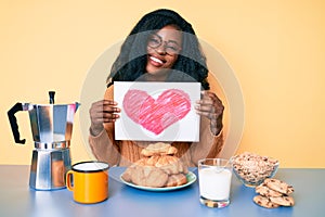 Beautiful african woman holding heart shape paper having breakfast looking positive and happy standing and smiling with a
