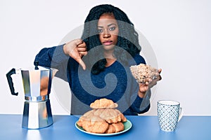 Beautiful african woman sitting on the table eating cereals with angry face, negative sign showing dislike with thumbs down,