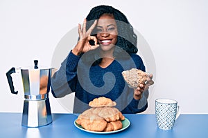 Beautiful african woman sitting on the table eating cereals doing ok sign with fingers, smiling friendly gesturing excellent