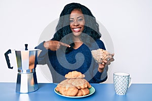 Beautiful african woman sitting on the table eating cereals pointing finger to one self smiling happy and proud