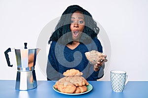 Beautiful african woman sitting on the table eating cereals scared and amazed with open mouth for surprise, disbelief face