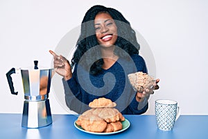 Beautiful african woman sitting on the table eating cereals smiling happy pointing with hand and finger to the side