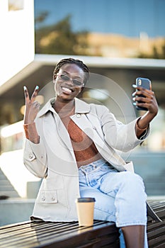 Beautiful African American woman taking selfie on mobile phone camera, showing victory sign. Emotional blogger streaming video