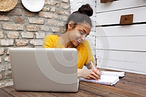 Beautiful african woman writing a book on cafe table with laptop