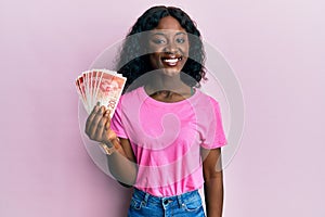 Beautiful african young woman holding 20 israel shekels banknotes looking positive and happy standing and smiling with a confident