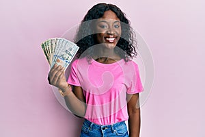 Beautiful african young woman holding dollars looking positive and happy standing and smiling with a confident smile showing teeth