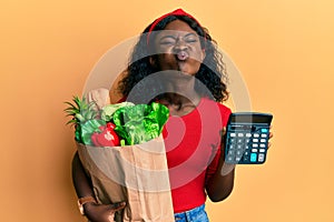 Beautiful african young woman holding groceries and calculator puffing cheeks with funny face