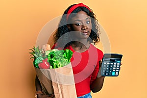 Beautiful african young woman holding groceries and calculator relaxed with serious expression on face