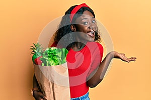 Beautiful african young woman holding paper bag with groceries celebrating achievement with happy smile and winner expression with