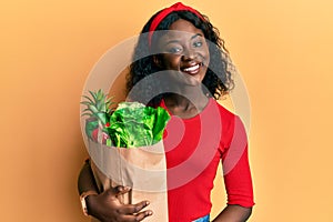 Beautiful african young woman holding paper bag with groceries looking positive and happy standing and smiling with a confident