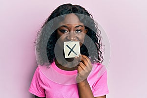 Beautiful african young woman holding paper with cross symbol looking positive and happy standing and smiling with a confident