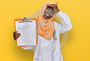 Beautiful african young woman wearing doctor stethoscope holding clipboard stressed and frustrated with hand on head, surprised