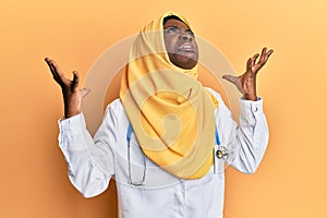 Beautiful african young woman wearing doctor uniform and hijab crazy and mad shouting and yelling with aggressive expression and