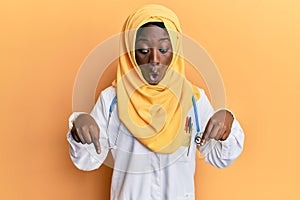 Beautiful african young woman wearing doctor uniform and hijab pointing down with fingers showing advertisement, surprised face