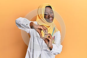 Beautiful african young woman wearing doctor uniform and hijab smiling in love doing heart symbol shape with hands
