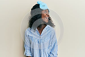 Beautiful african young woman wearing sleep mask and pajama looking away to side with smile on face, natural expression