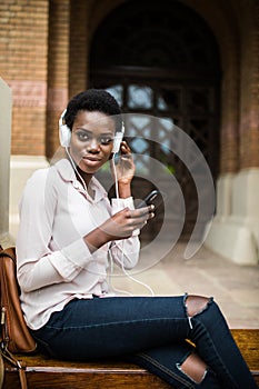 Beautiful afro american girl listening to music through headphones sitting on a bench. female with afro hair watching video on sma