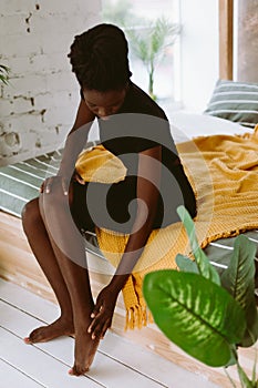 Beautiful afro american woman sitting on bed in decorated studio and massaging foot by hand. Black person having sports