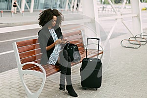 The beautiful afro-american woman is sitting on the bench and chatting via the mobile phone near the airport.