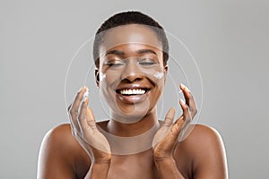 Beautiful afro woman applying cream on her cheeks and sincerely smiling