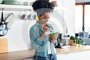 Beautiful afro woman eating noodles with chopsticks while standing in the kitchen at home