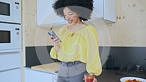 Beautiful afro woman sending messages with her smartphone while drinking tea in the kitchen at coworking place.