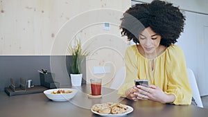 Beautiful afro woman sending messages with her smartphone while drinking tea in the kitchen at coworking place.