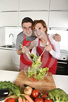 Beautiful American couple working at home kitchen in apron mixing vegetable salad smiling happy