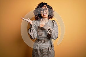 Beautiful arab business woman wearing dress and glasses standing over yellow background Showing palm hand and doing ok gesture