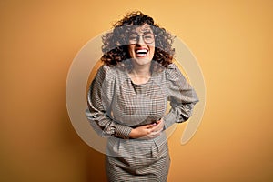 Beautiful arab business woman wearing dress and glasses standing over yellow background smiling and laughing hard out loud because