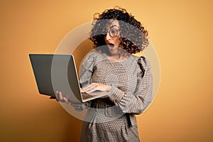 Beautiful arab businesswoman wearing glasses working using laptop over yellow background scared in shock with a surprise face,