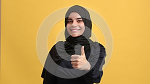 Beautiful Arab woman in black hijab smiling showing cool thumb up gesture posing isolated on orange