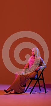 Beautiful arab woman posing in stylish hijab isolated on brown studio background in neon light. Fashion, beauty, style